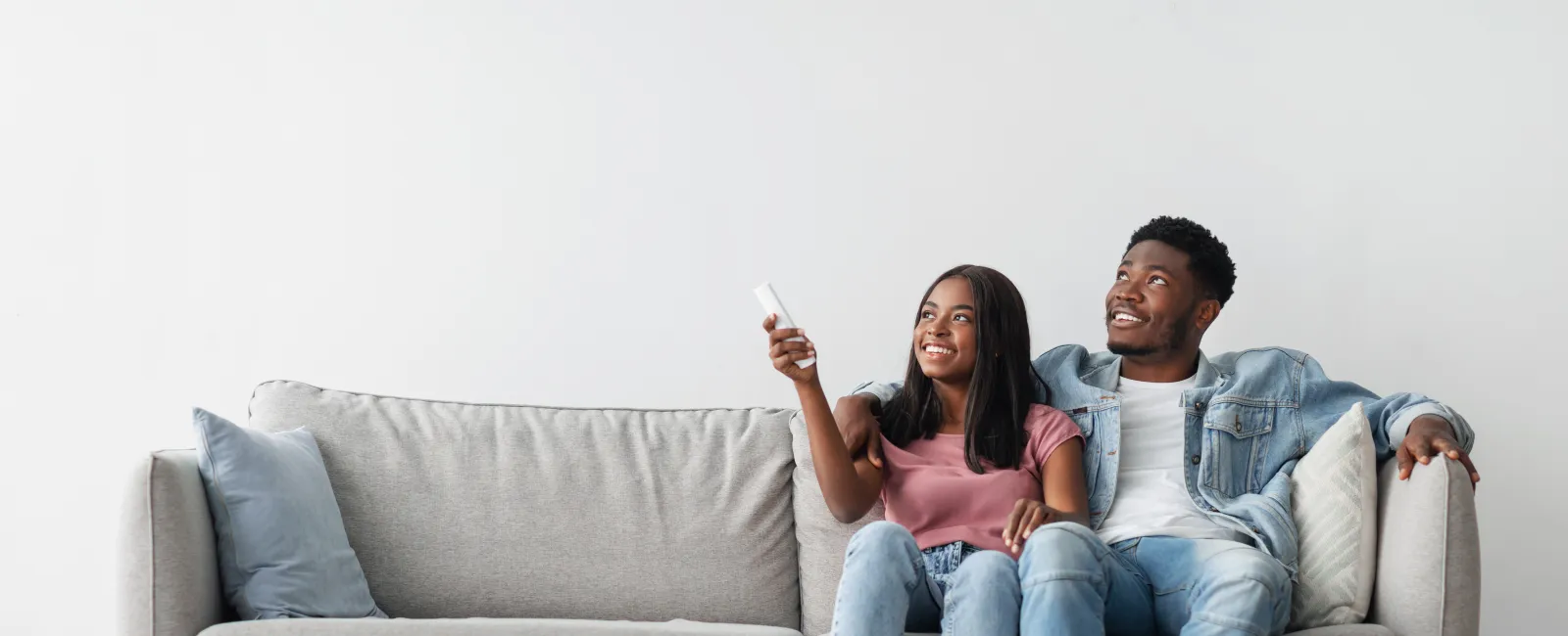 Young couple sitting on a gray couch smiling and holding a remote in a bright living room