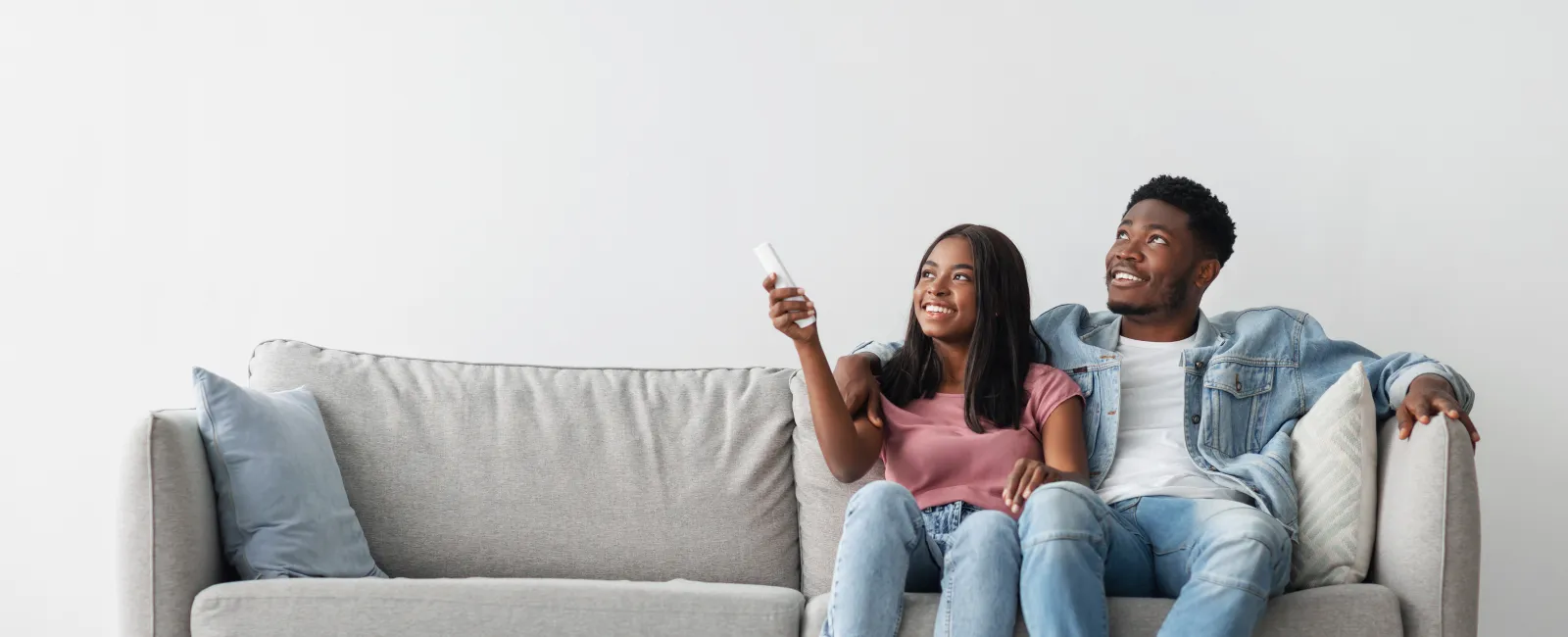 Happy young couple sitting on modern gray sofa watching TV with remote control in hand