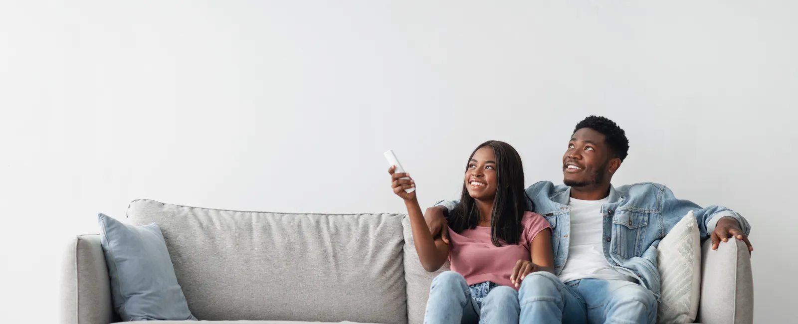 Young couple sitting on gray couch smiling, woman holding remote control, looking up enjoy television time