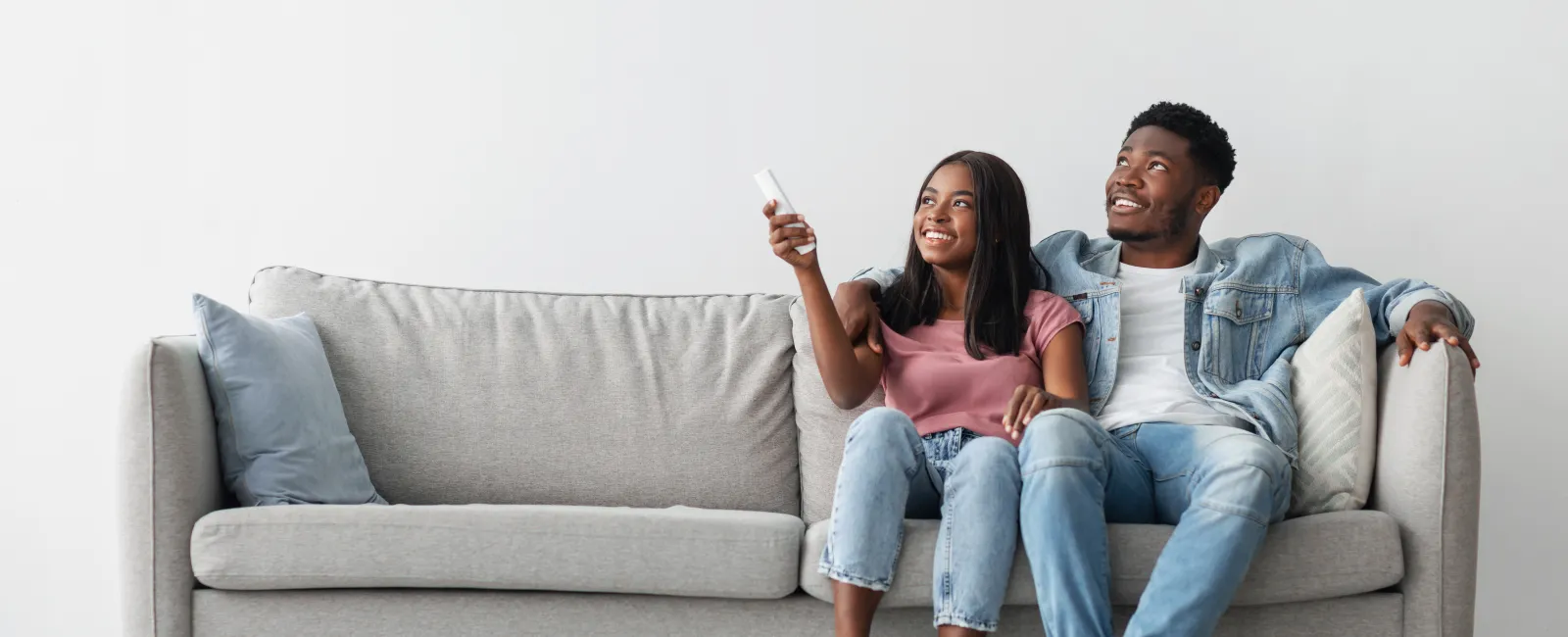 Happy young couple sitting on gray sofa watching TV with remote in modern living room