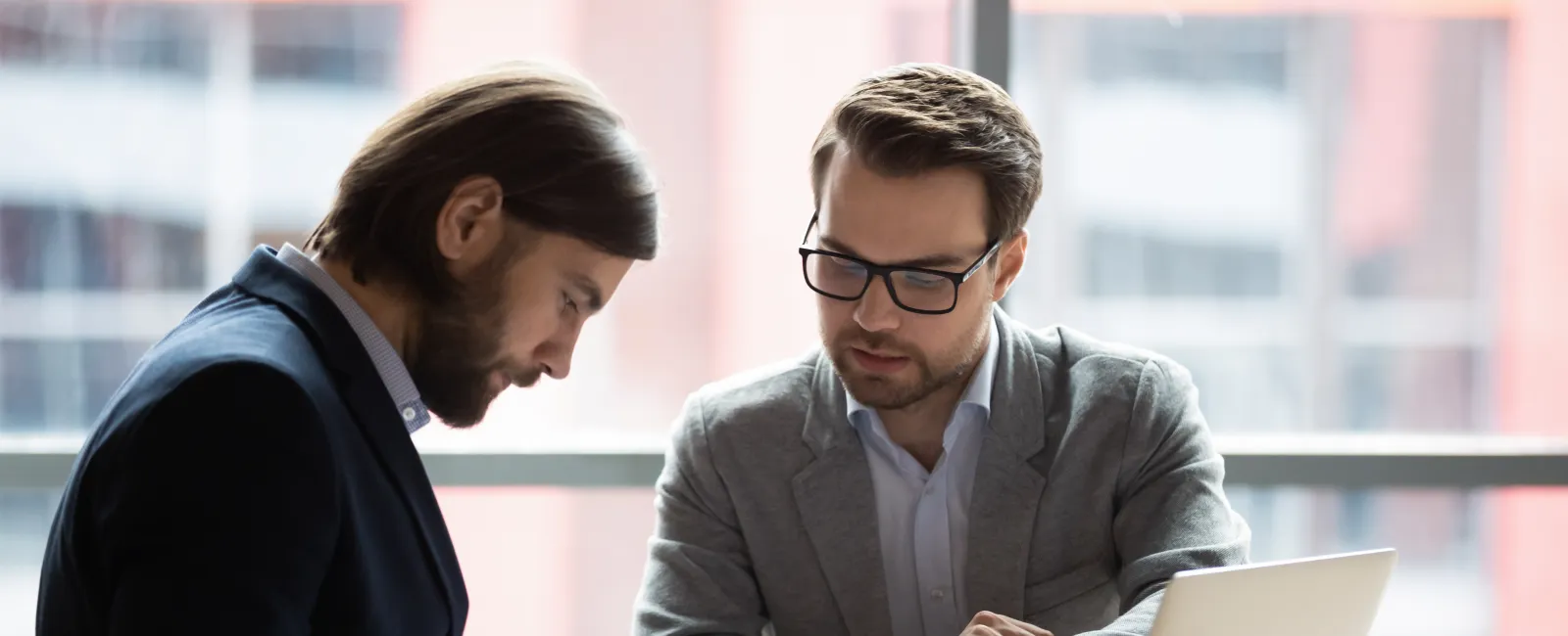 Two businessmen in office discussing documents with laptop, reviewing paperwork and collaborating on project.