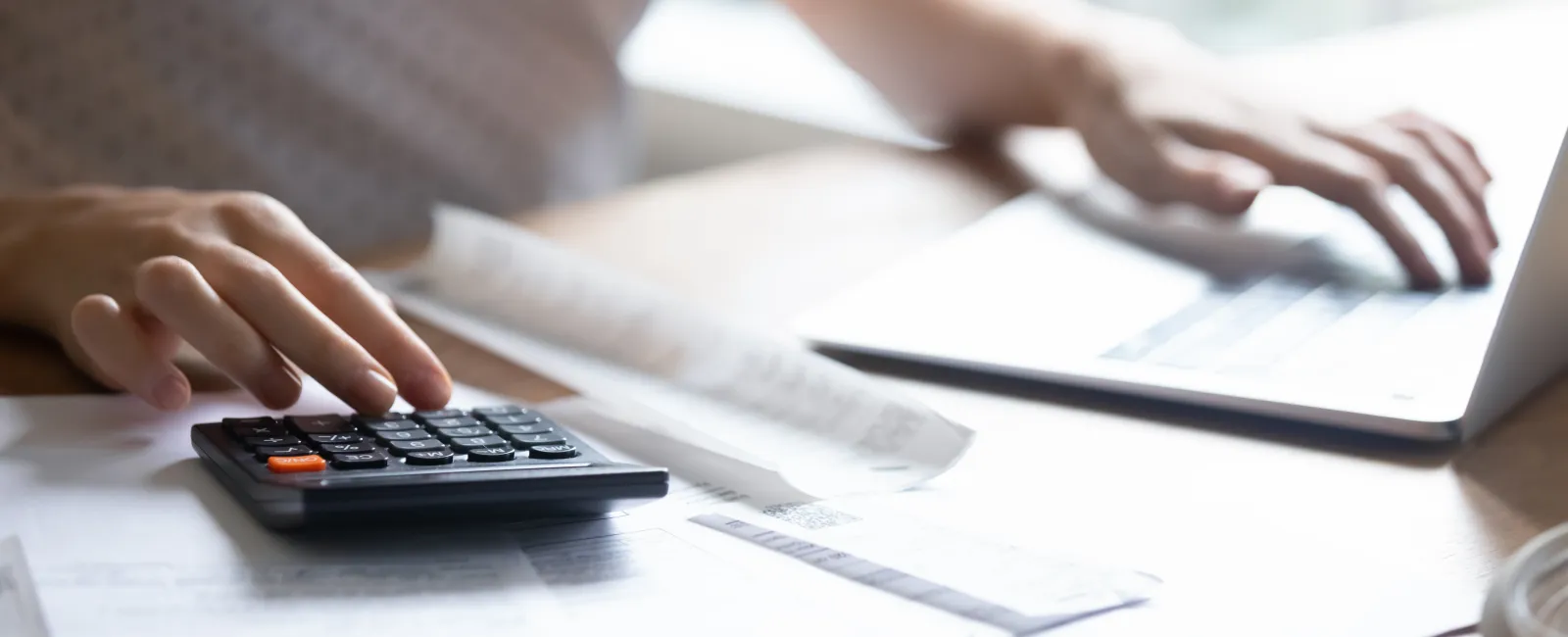 Person calculating bills with calculator and using laptop on wooden table with papers and receipts.