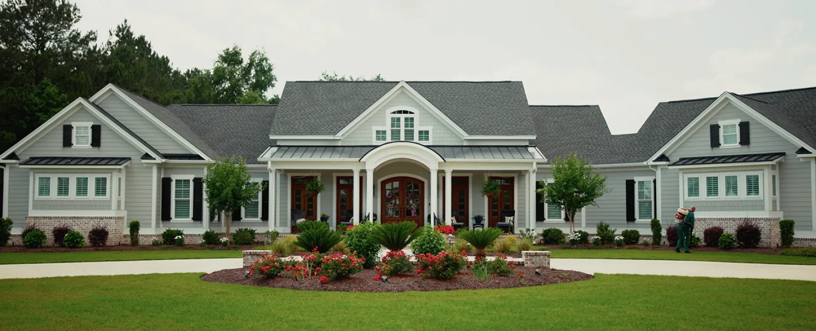 Spacious white house with gray roof, large front porch, lush lawn, and landscaped garden with flowers and shrubs.