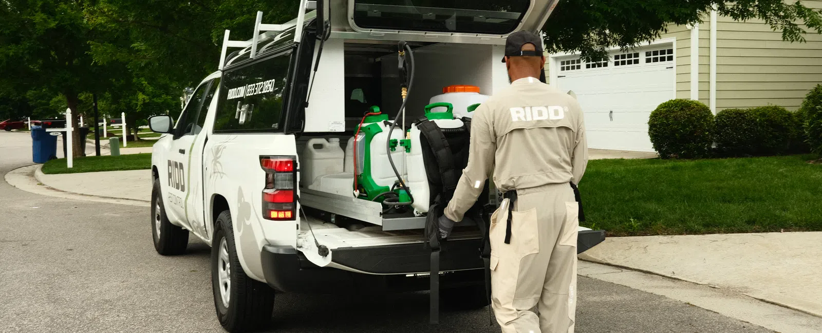 Pest control worker loading equipment into a white RIDO service truck parked in a residential neighborhood.