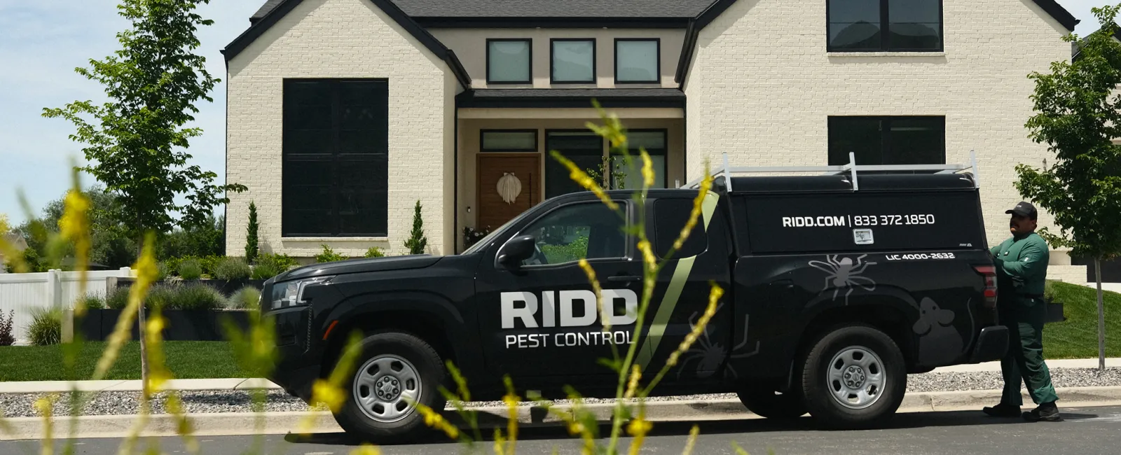 Black RIDDO Pest Control truck parked outside a modern suburban house with technician standing nearby.