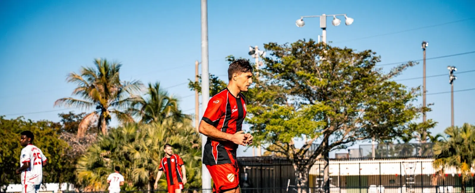 Soccer player in red and black uniform running on field during daytime match under clear blue sky.