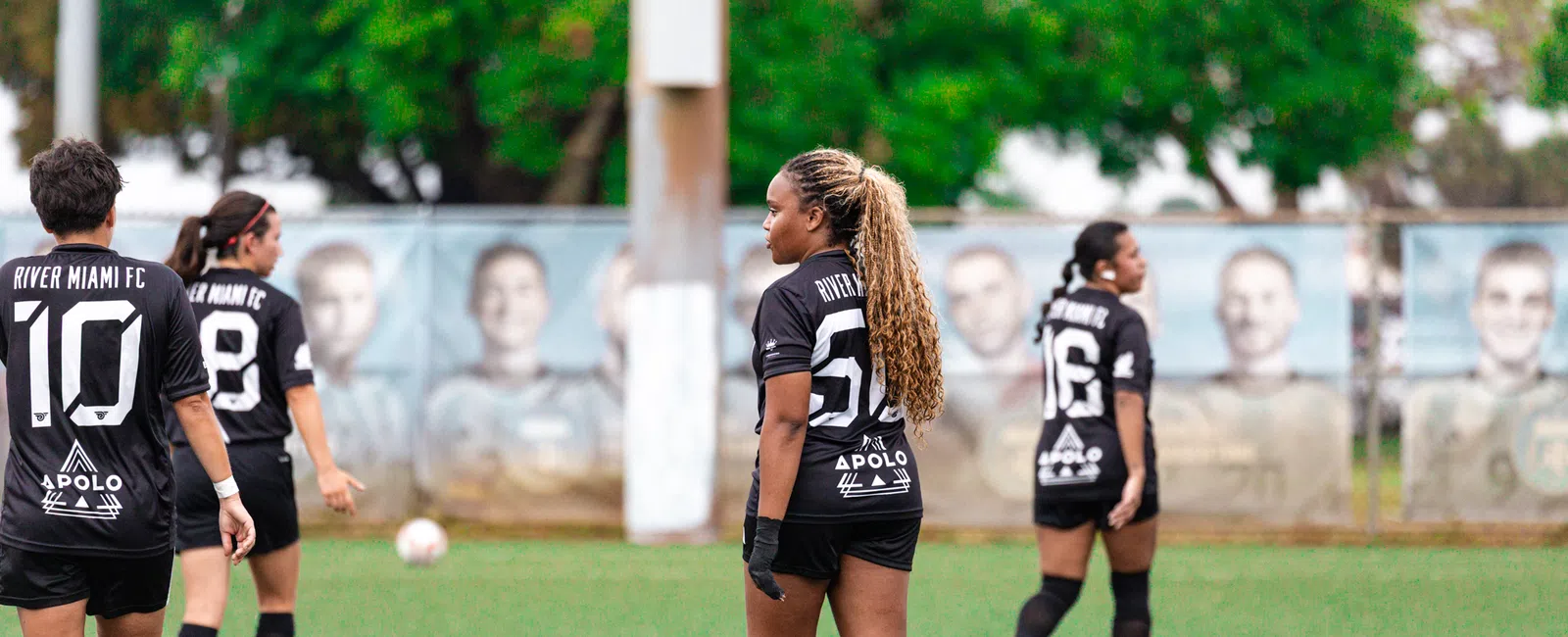 Women soccer players in black River Miami FC uniforms on a green field during a game or practice session.