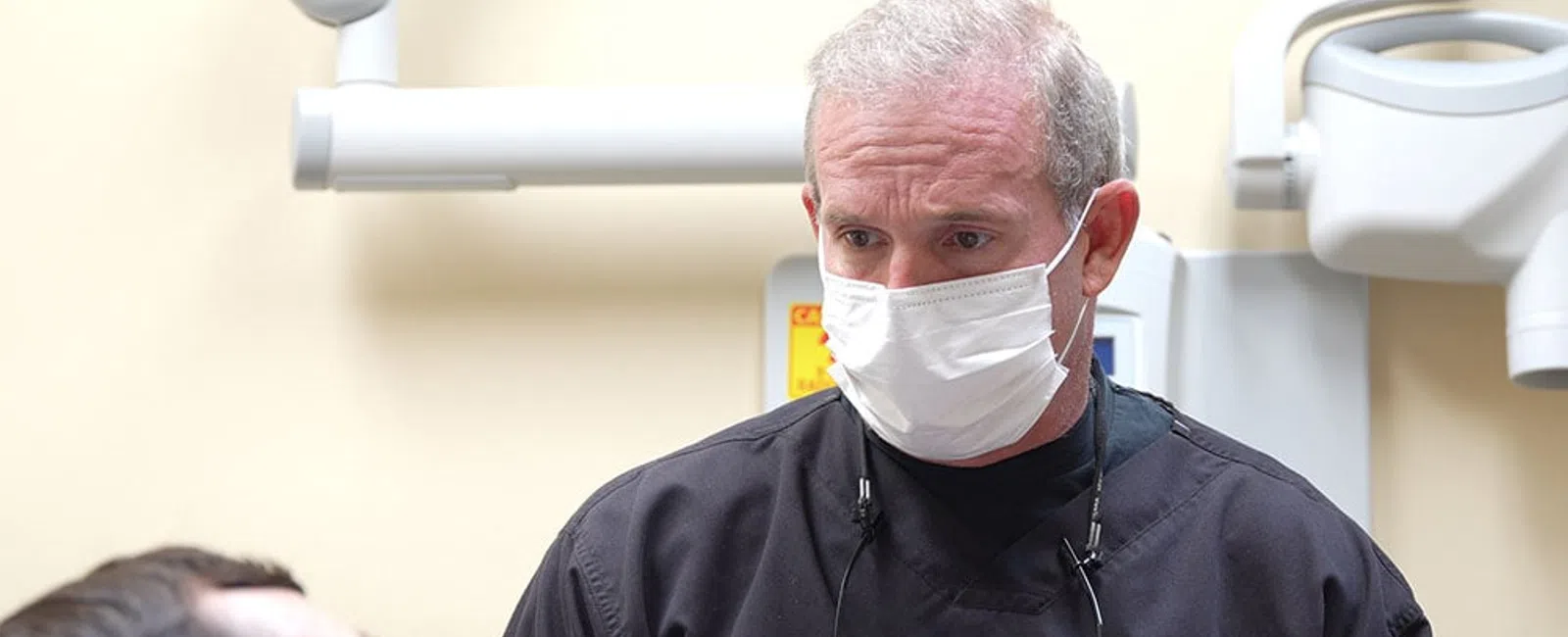 Dentist wearing a mask and black scrubs examining a patient in a dental clinic with equipment in the background