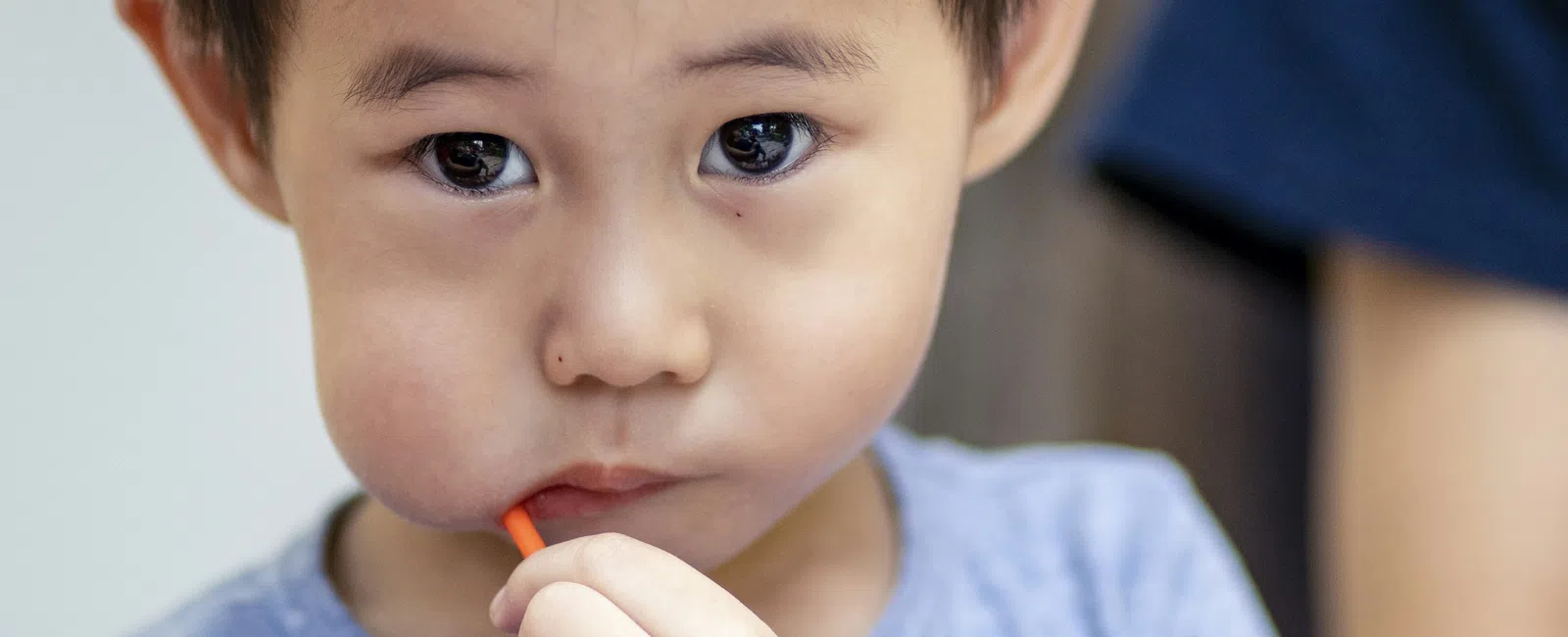 Young boy with dark hair in blue shirt sucking on a red straw, looking attentively at the camera indoors.
