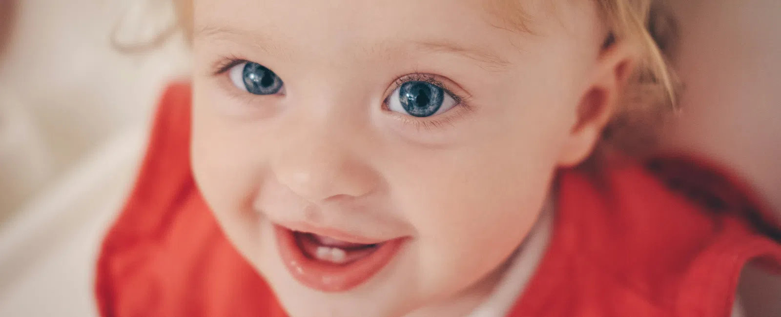 Close-up of a smiling baby with blue eyes and blonde hair wearing a red outfit, looking happily at the camera