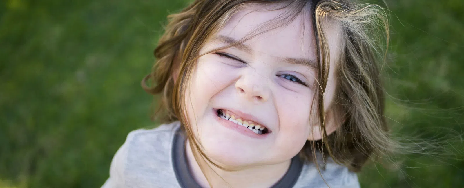 Smiling young child with tousled hair wearing a gray shirt outdoors on green grass background