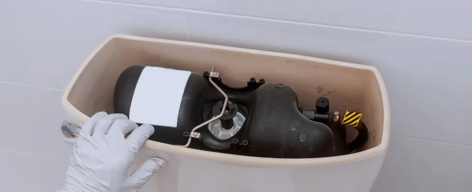 Person wearing gloves inspecting the inside of a toilet tank with visible components and plumbing parts.