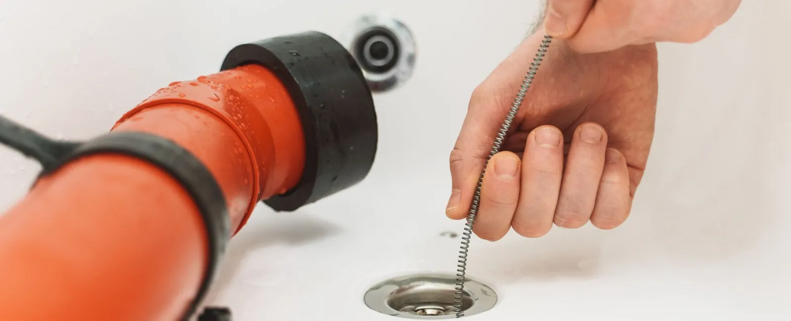 A person unclogging a sink with a plumbing snake near a red drainage pipe.
