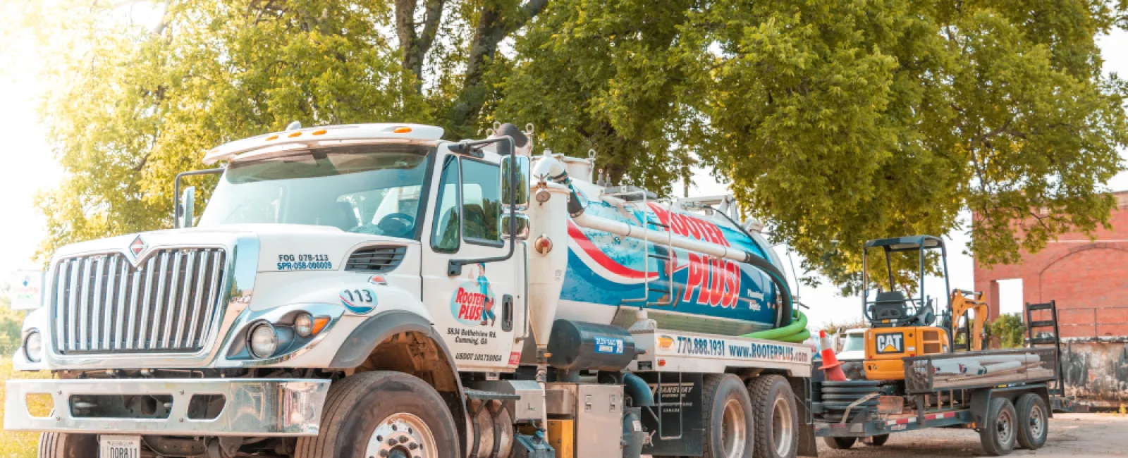 Rooder Plus septic service truck with tank and small excavator on trailer parked under green trees on sunny day.