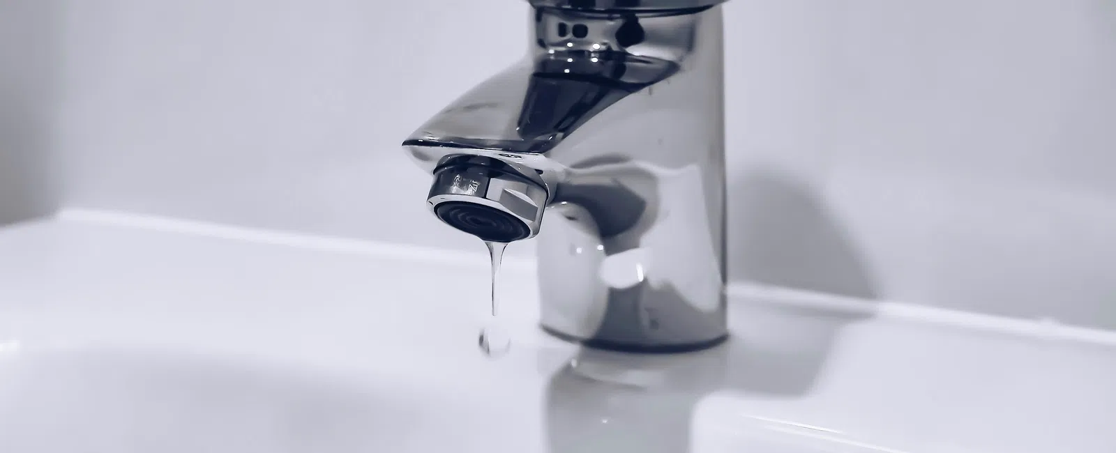 Chrome faucet leaking a drop of water above a white sink basin with drain hole.
