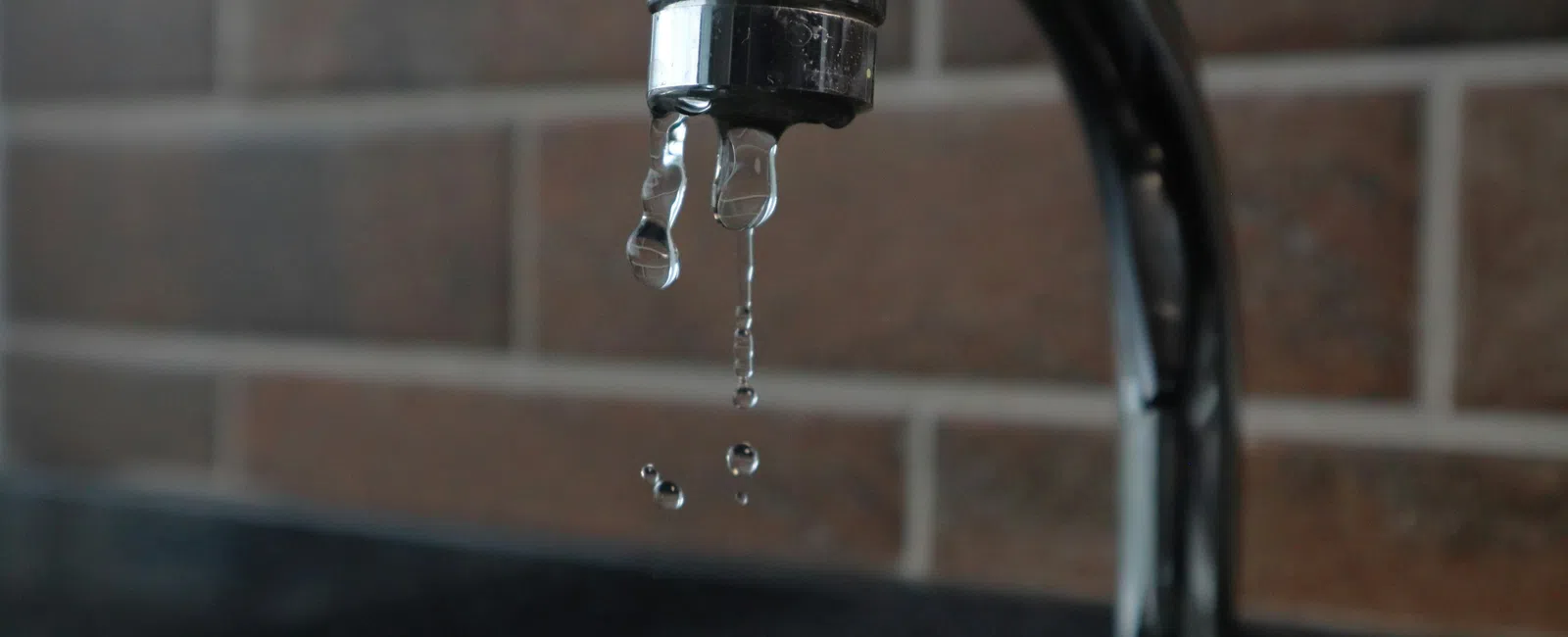Close-up of a dripping faucet with water droplets falling and brick wall background in a kitchen or bathroom.