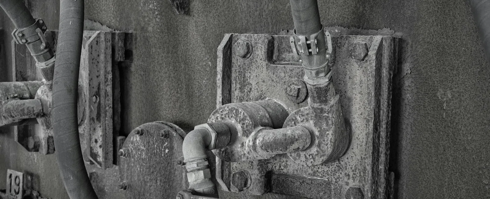 Close-up of rusted industrial pipes and connectors mounted on a weathered metal surface in black and white.