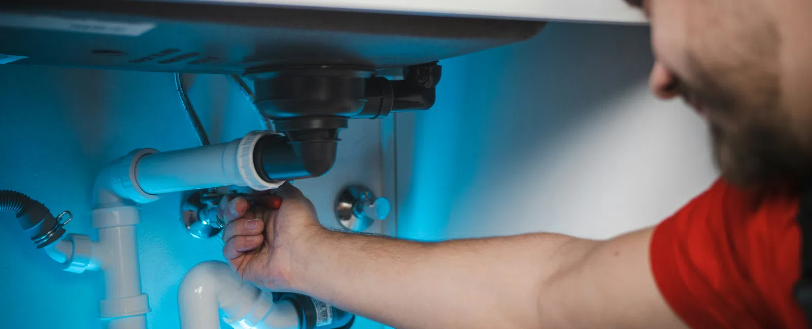 Man repairing white PVC pipes under a sink with blue lighting emphasizing plumbing work inside cabinet.