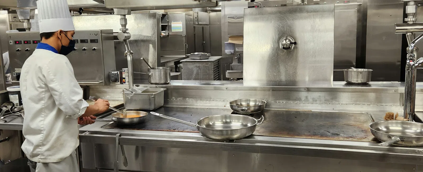 Chef in white uniform cooking with pans on a large stainless steel stovetop in a commercial kitchen.