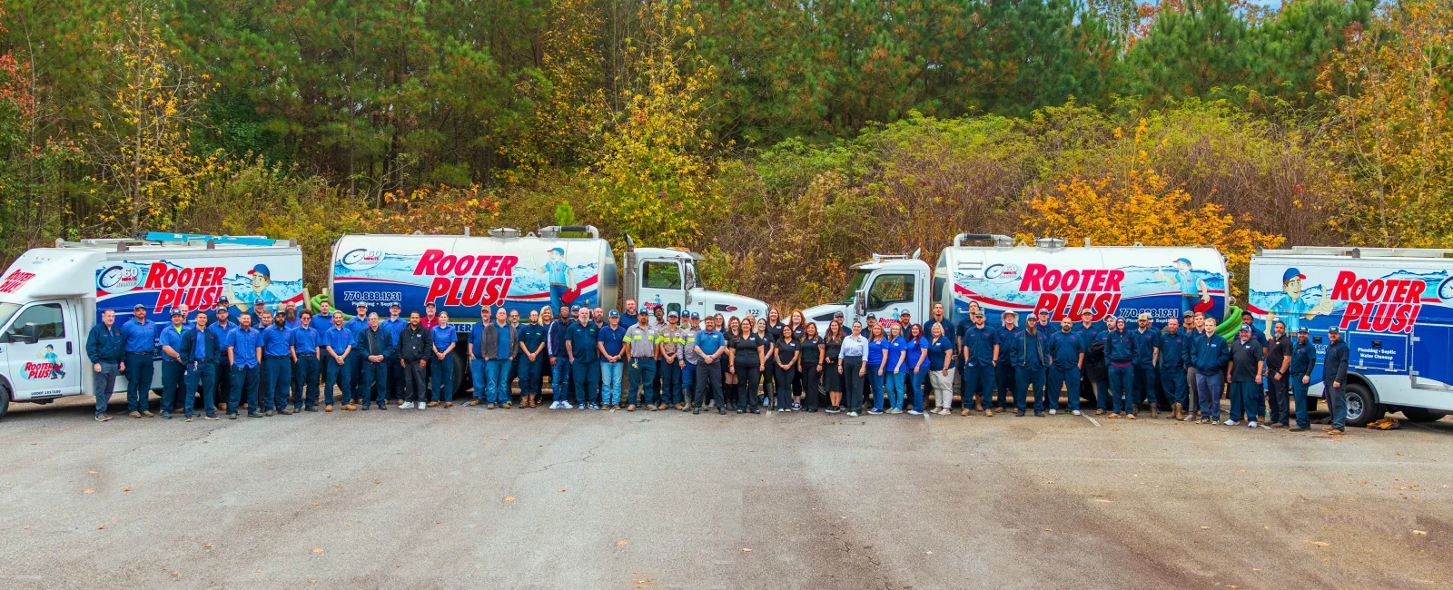 Large Rooter Plus! plumbing team standing in front of five service trucks outdoors with trees in fall colors behind them.