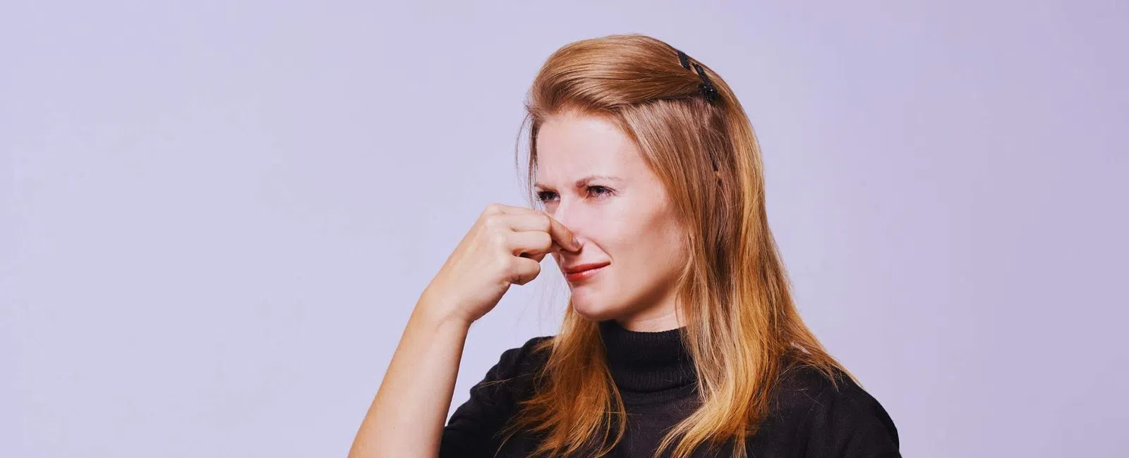 Woman in black sweater pinching her nose with a disgusted expression against a light purple background