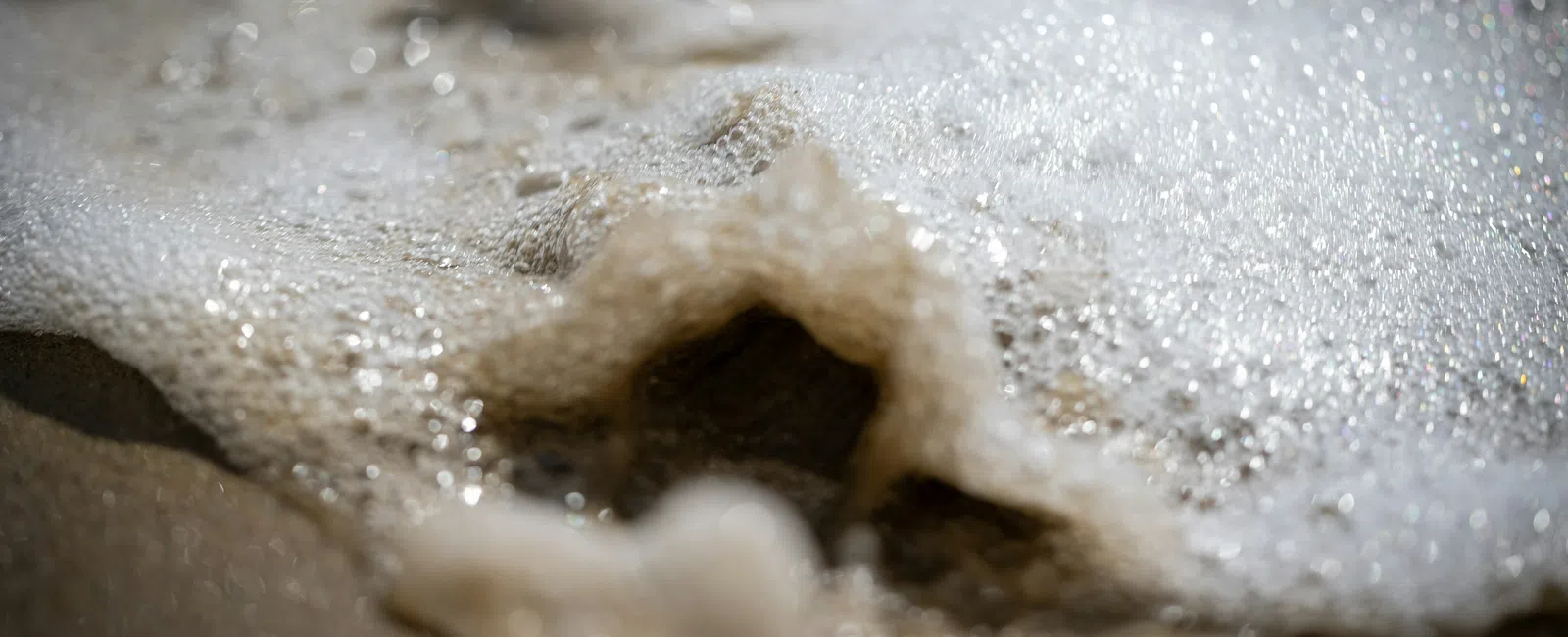 Close-up of sea foam bubbles on wet sandy beach with soft sunlight reflecting on water surface.
