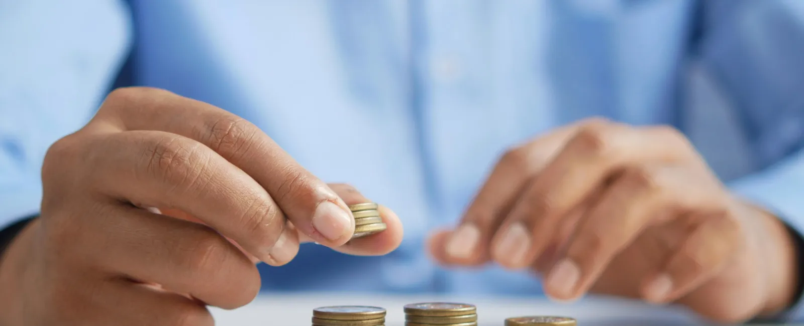 Person stacking coins into three small piles on a white surface with a blurred blue shirt background.