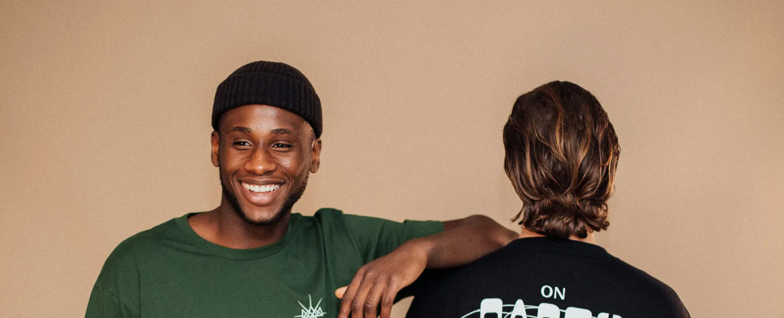 Two men modeling casual streetwear with printed designs, one smiling in green tee, other showing text on black sweatshirt.