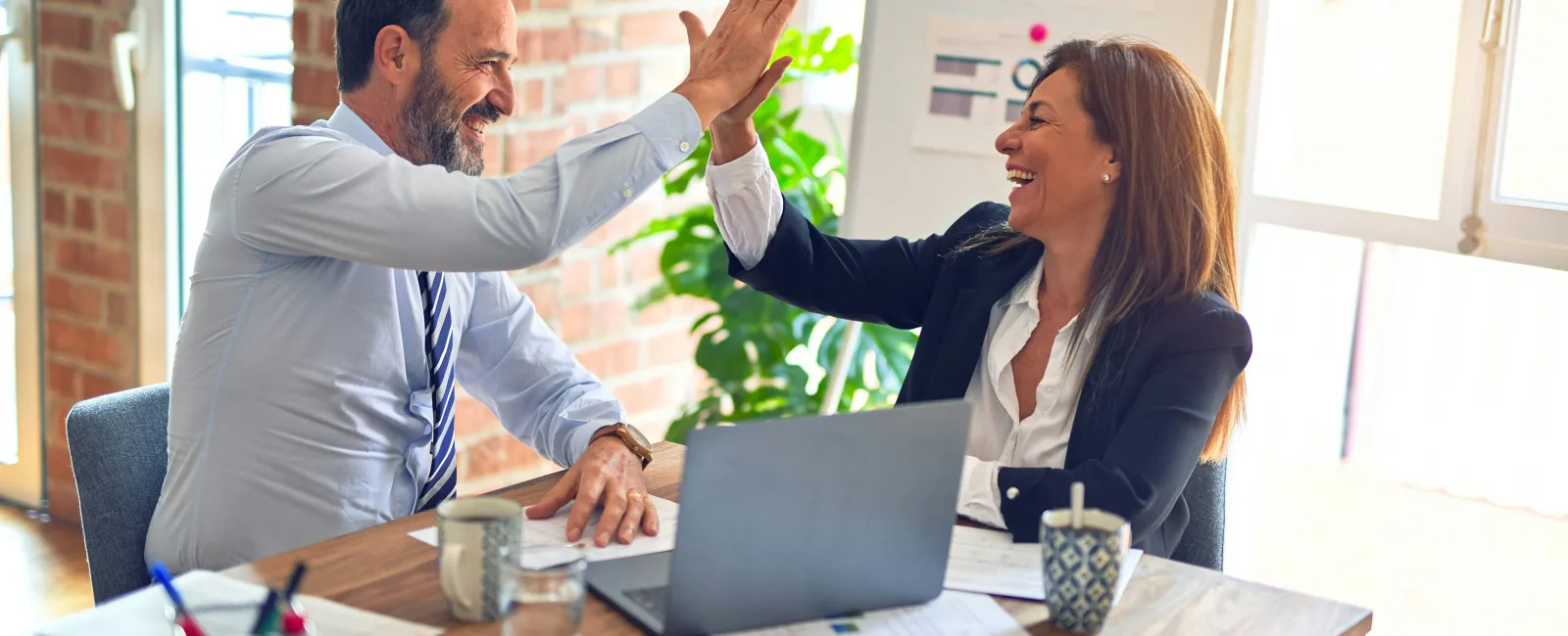 Business colleagues in office celebrating success with a high five over a desk with laptop and documents.