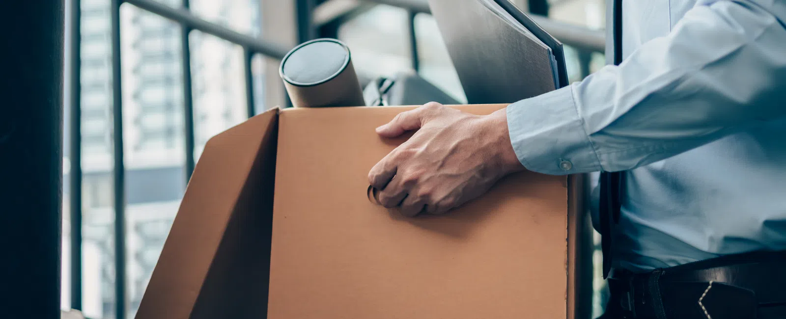 Businessperson carrying a cardboard box with office items outside a modern office building, symbolizing job change or moving.