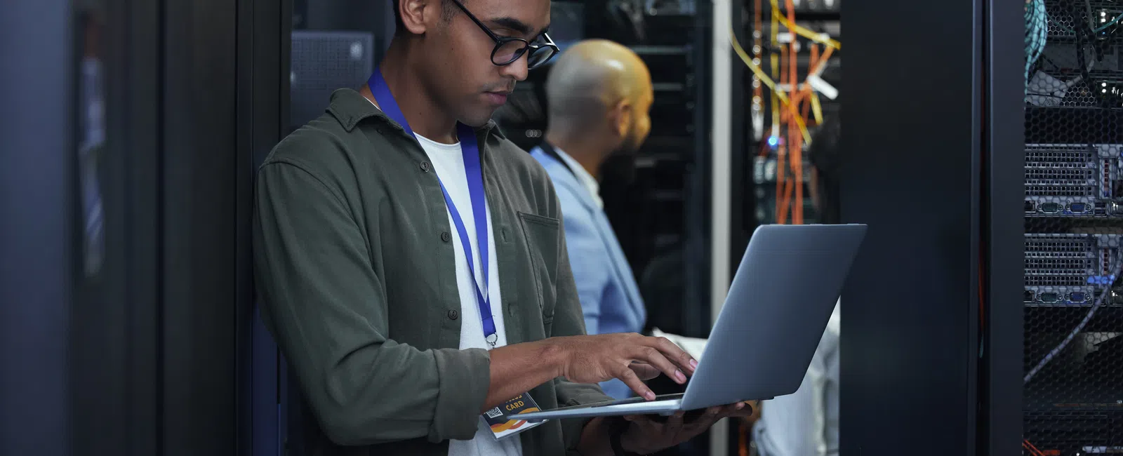 IT technician working on a laptop in a server room surrounded by networking equipment and cables