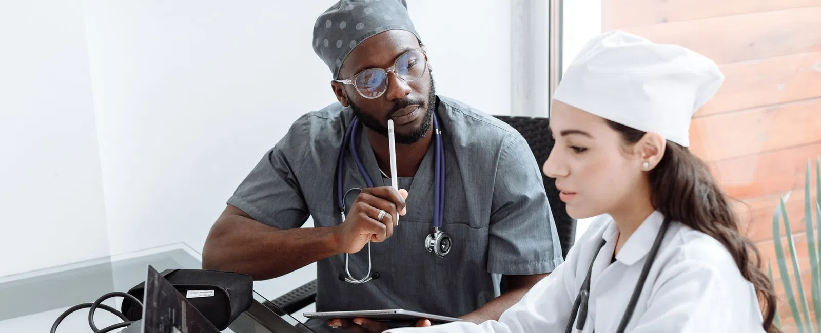 Two doctors in scrubs and lab coats collaborating over a laptop in a modern medical office with stethoscopes.