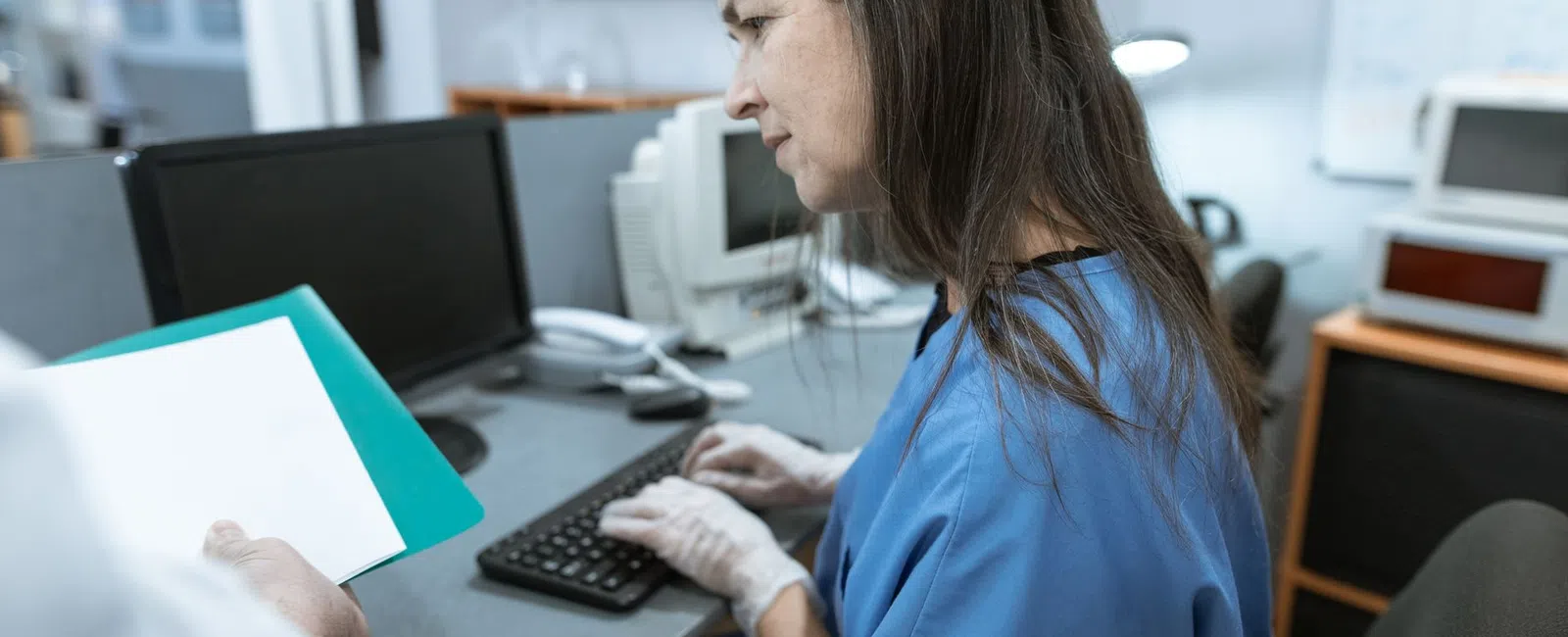 Medical professional in blue scrubs working at computer with patient file in clinical office setting.