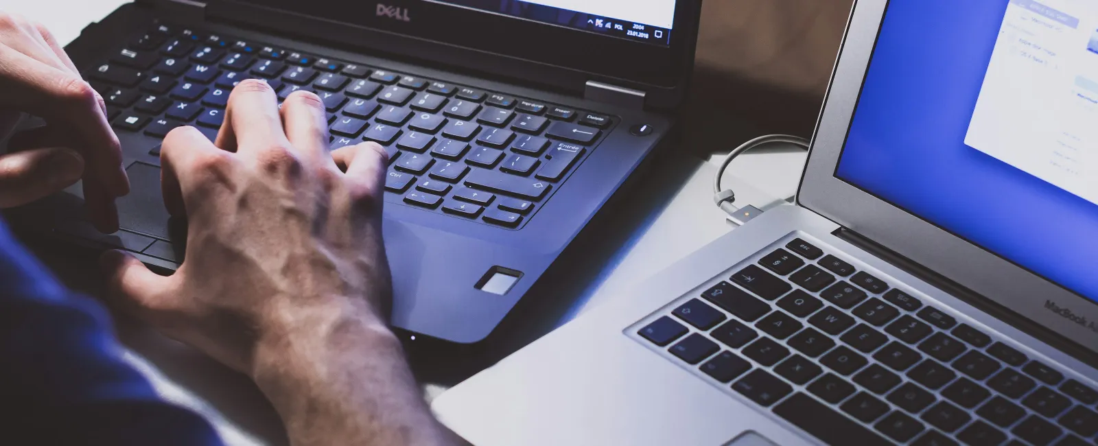 Person typing on a Dell laptop next to an open MacBook Air on a white desk in a workspace.
