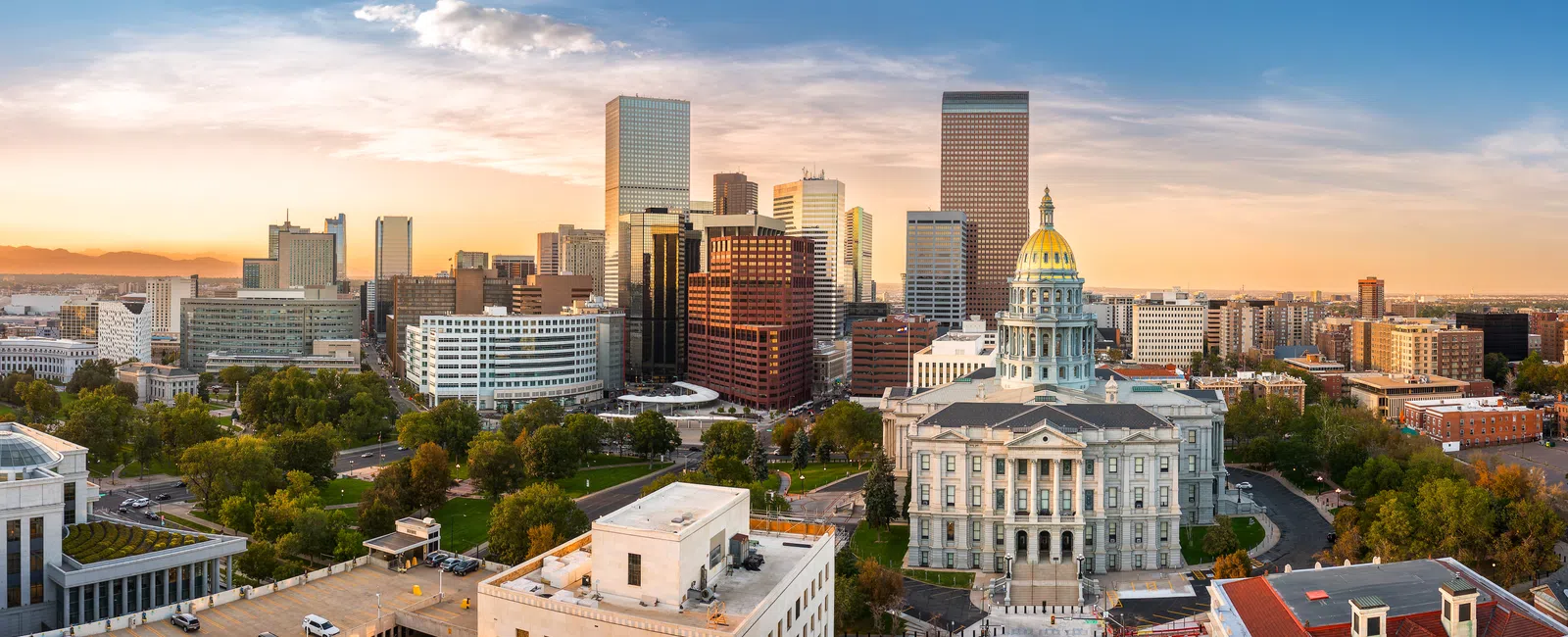 Denver city skyline at sunset with Colorado State Capitol and modern skyscrapers amid green trees.