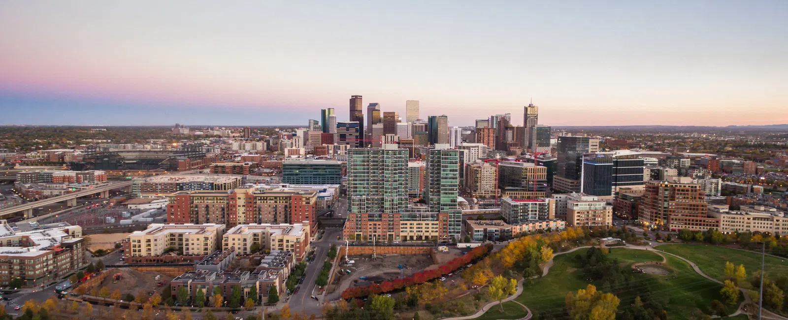 Aerial view of downtown city skyline with modern buildings and green parkland under a clear sky at sunset.