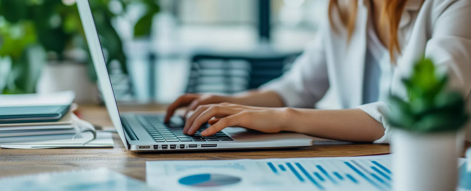 Young professional woman working on laptop at desk with charts, papers, and plants in modern office.