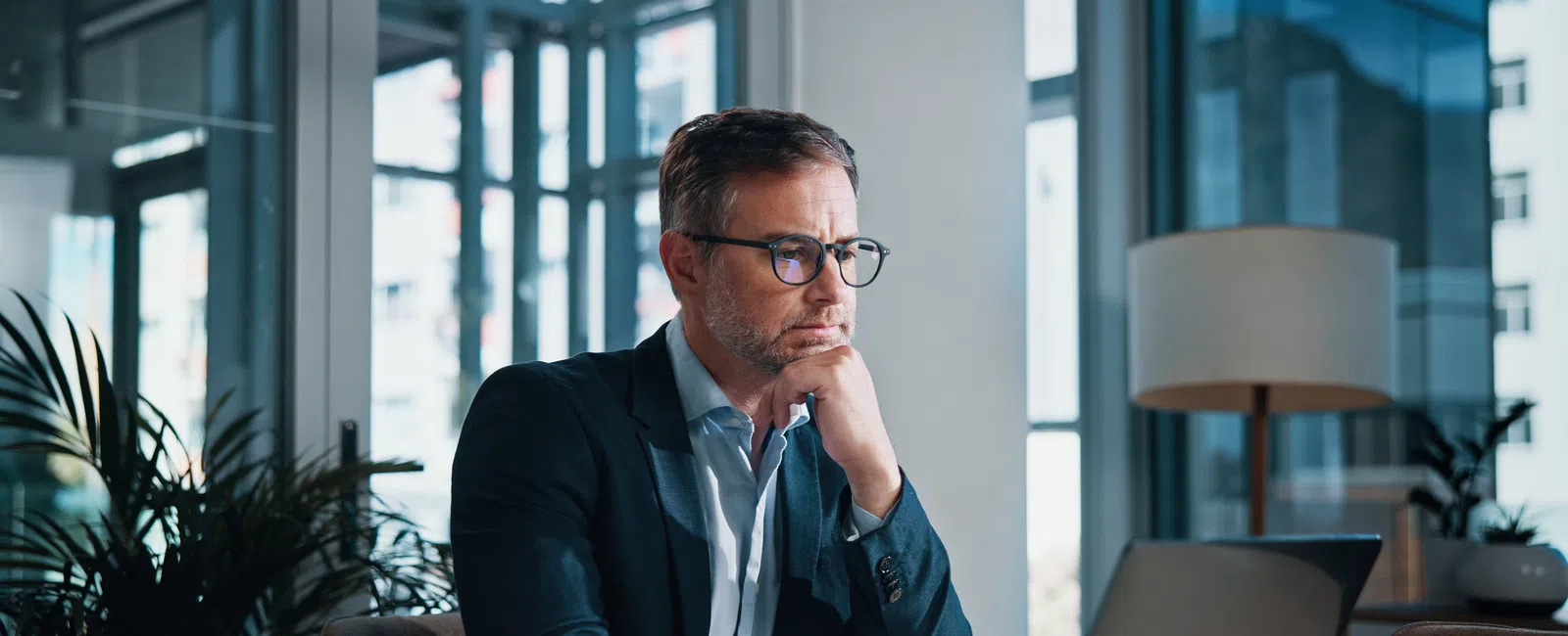 Focused businessman wearing glasses working on laptop in modern office with large windows and plants.