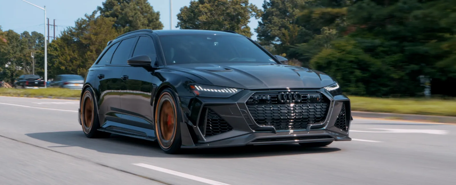Black Audi RS6 Avant driving on a road with bronze rims and trees in the background under clear sky.