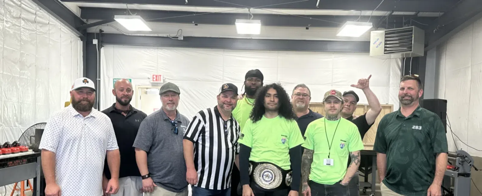 Group of men standing in a workshop, one wearing a championship belt, others in casual and referee attire.