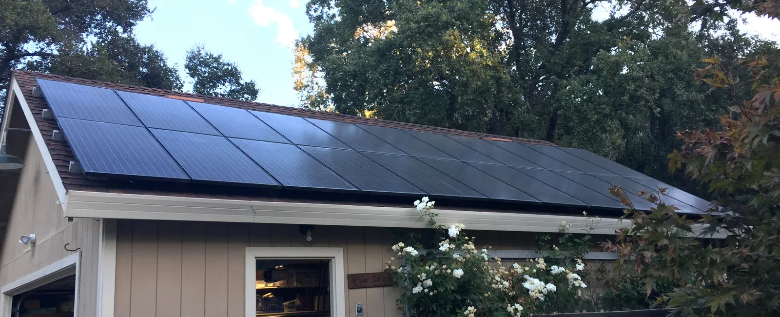 Residential roof with black solar panels installed amid surrounding trees under a clear sky.