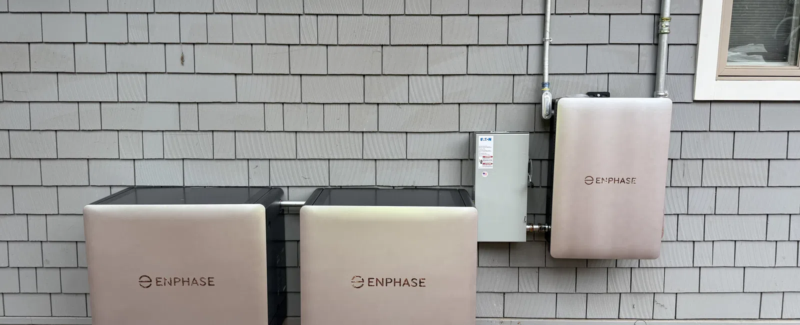 Three Enphase energy storage units mounted on a gray shingled exterior house wall under two windows.
