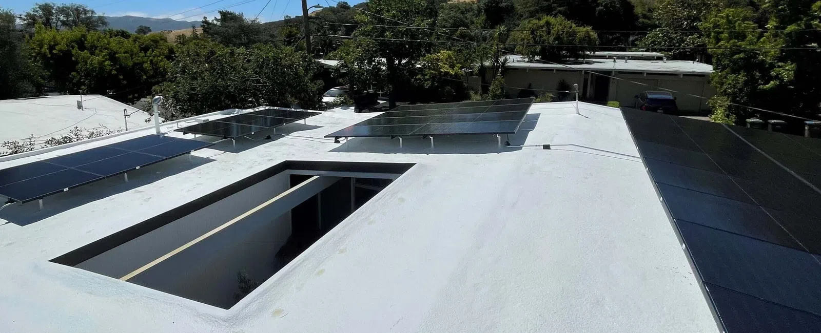 Solar panels installed on a white flat rooftop with trees and mountains in the background on a sunny day