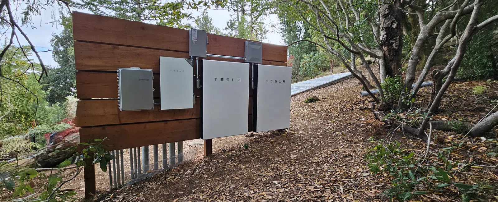 Tesla Powerwall home battery units installed on a wooden panel in an outdoor wooded area.