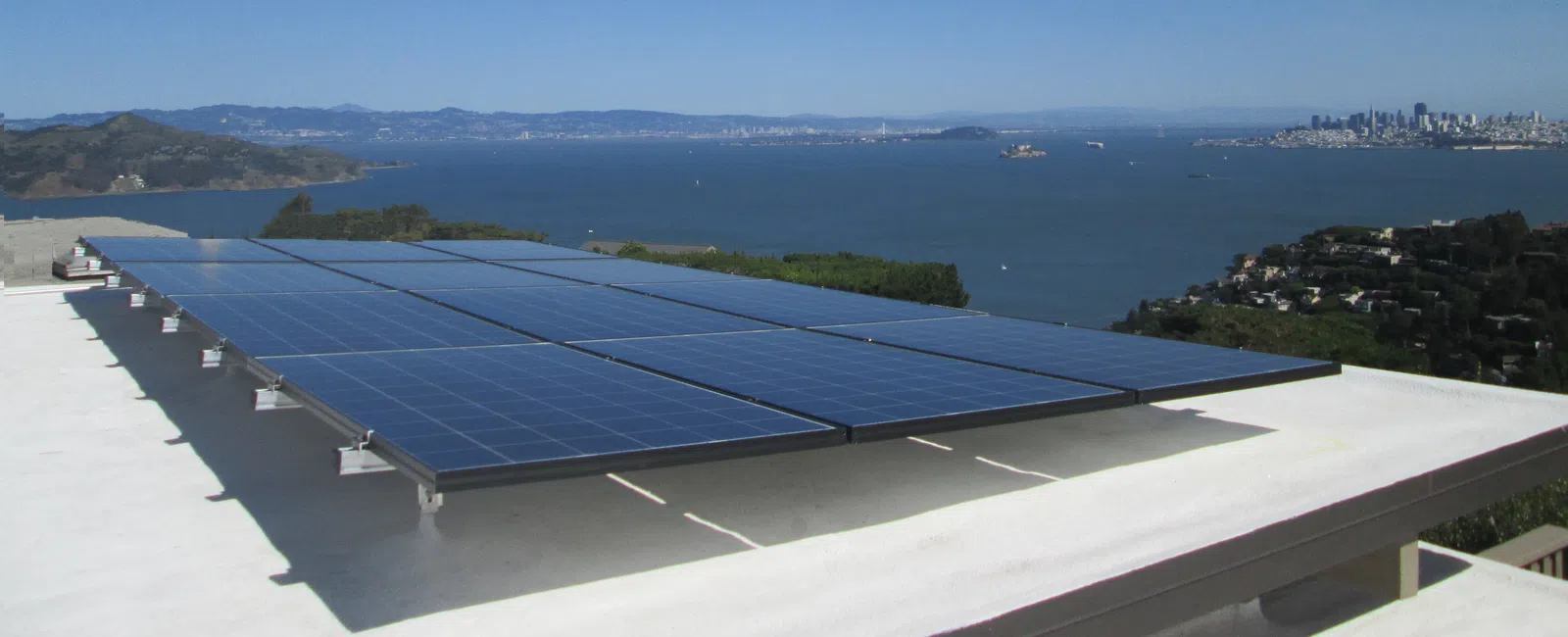 Solar panels installed on a rooftop overlooking a bay with a city skyline in the background under a clear blue sky.