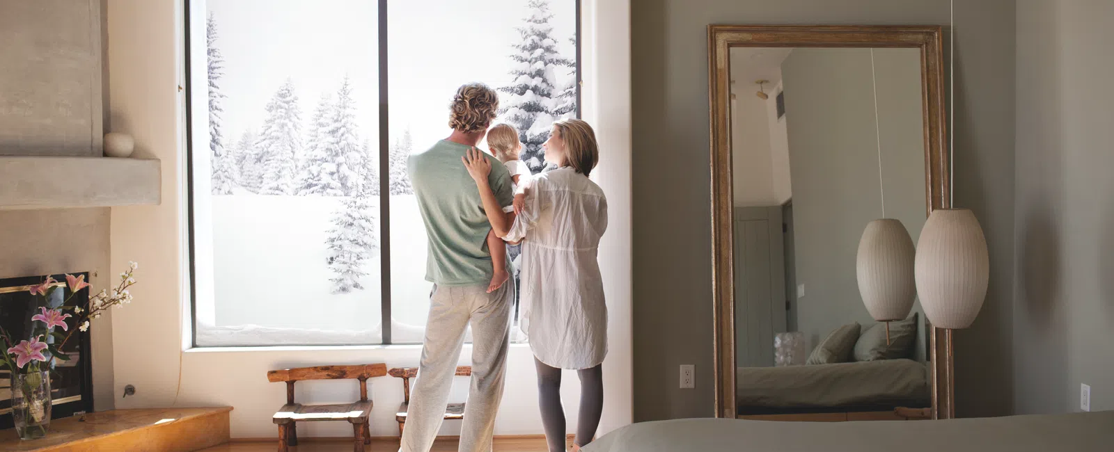 Family of three standing in a cozy bedroom looking out a large window at snowy trees outside during daytime.