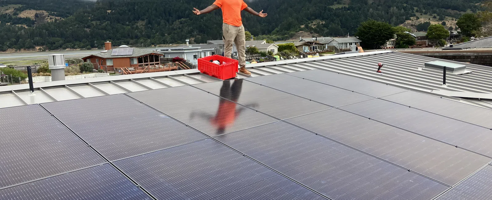 Person in orange shirt standing on a roof with solar panels under a cloudy sky.
