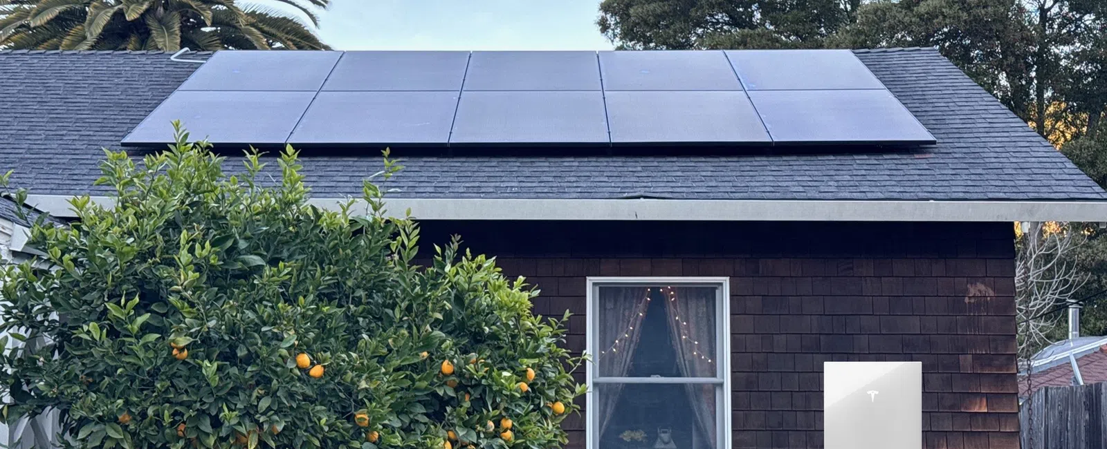 Solar panels on a house roof with a Tesla Powerwall battery mounted on the exterior wall beside a window and orange tree.