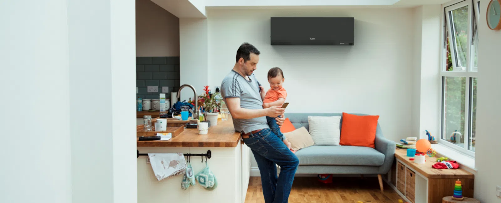 Father and child using smartphone in bright living room with sofa, air conditioner, and toys on wooden floor.