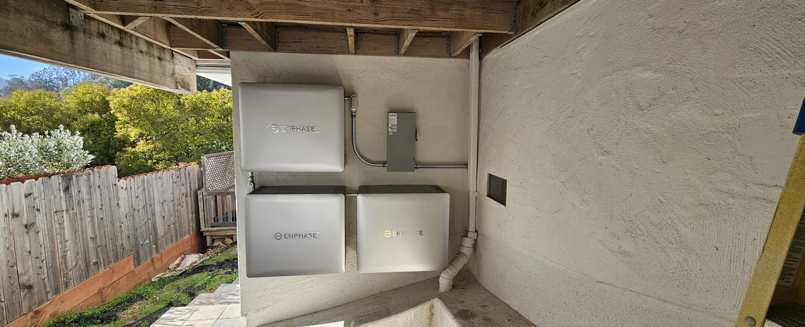 Three Enphase energy storage batteries mounted on an outdoor wall under a wooden roof near a fenced yard.